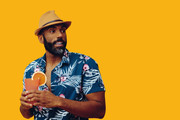 handsome african american man wearing hawaian shirt and hat with orange juice posing on yellow background - Фото, изображение