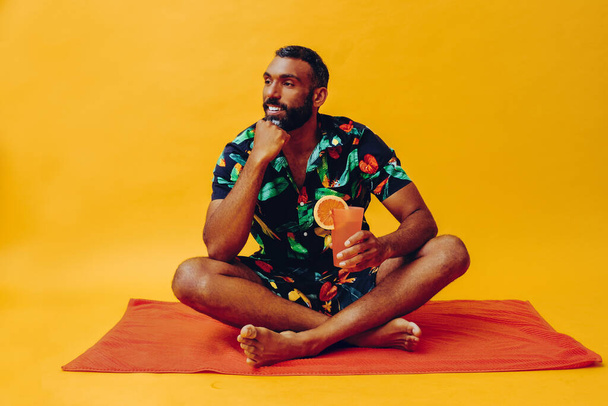 handsome african american man on vacation sitting on an orange towel, holding orange juice - Φωτογραφία, εικόνα