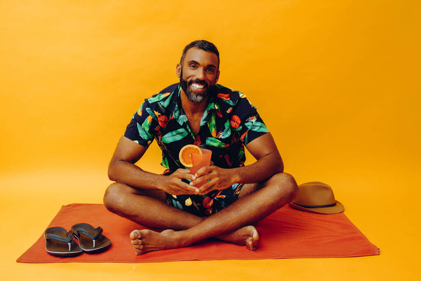 handsome african american man on vacation sitting on an orange towel, holding orange juice - Foto, Bild
