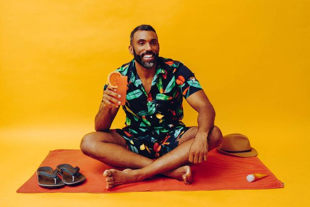 handsome african american man on vacation sitting on an orange towel, holding orange juice - Foto, imagen