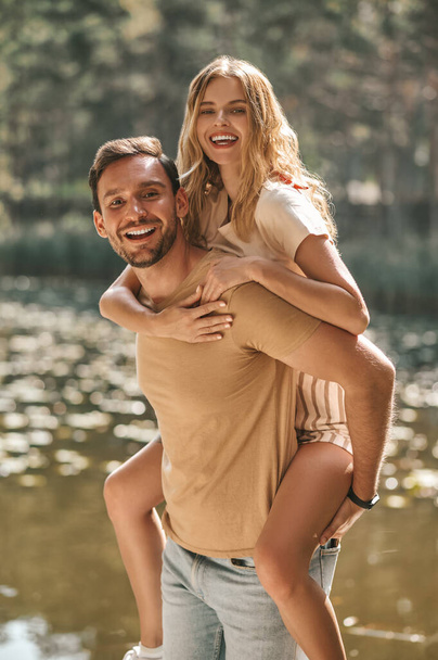 Smiling strong energetic romantic male carrying a happy attractive young blonde woman on his back - Foto, afbeelding