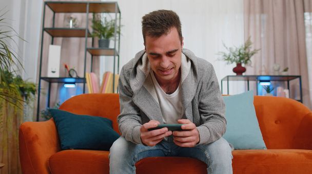 Беспокойный юный взрослый мужчина с энтузиазмом играет в шутеры онлайн видеоигры на смартфоне. Возбужденный мальчик игрок, использующий мобильный телефон приложение праздновать победу дома, сидя на диване в одиночку - Фото, изображение