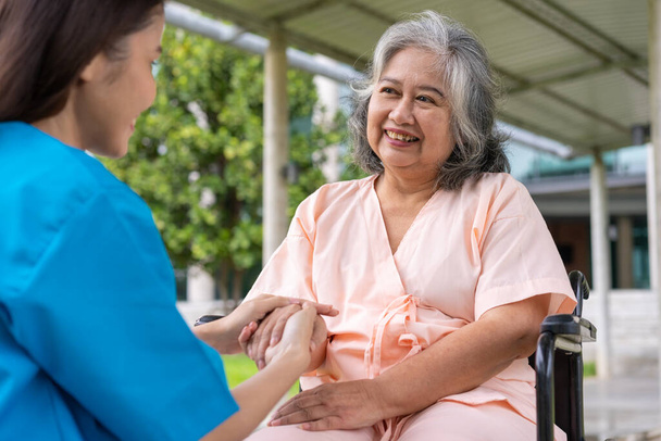 Asyalı dikkatli bakıcı ya da hemşire hastanın elini tutup hastayı tekerlekli sandalyeye oturtmaya teşvik ediyor. Bakıcının, birikimin ve sağlık sigortasının özenle mutlu emeklilik kavramı. - Fotoğraf, Görsel