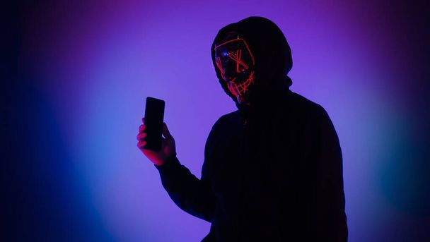 Névtelen hacker és maszk okostelefonnal a kezében. Egy fekete csuklyás férfi próbál személyes adatokat meghackelni a mobiljáról. Számítógépes bűnözés adatainak hackelése vagy személyes adatok ellopása koncepció képviselete. - Fotó, kép