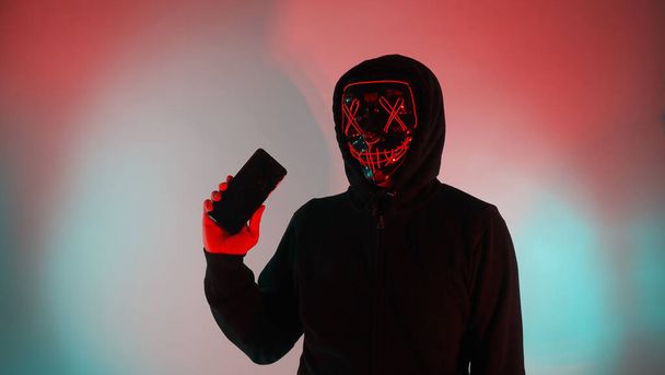 Hackeador anónimo y máscara facial con smartphone en la mano. Hombre con camisa de capucha negra tratando de hackear datos personales desde el teléfono móvil. Representar el concepto de piratería informática o robo de datos personales. - Foto, imagen