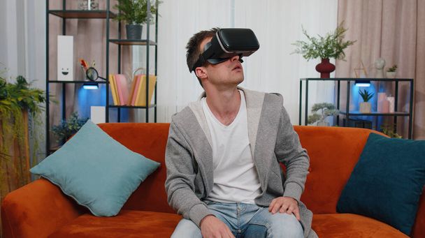 Surprised man use VR headset glasses at home, enjoying 3D 360 video game concept, entertaining, in positive mood, moving hands in air, sitting in living room. Virtual reality, modern gadget device - Fotografie, Obrázek