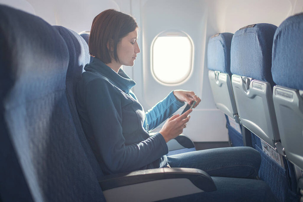 Young beautiful woman sitting at window of plane during the flight. reading a book on a smartphone, playing apps, writing notes - Photo, Image