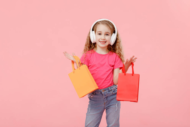 adorable child girl in headphones is holding shopping bags in hands above head isolated on pink background. shopping concept. - Foto, immagini