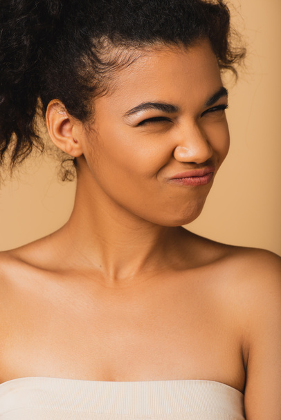 curly african american woman with bare shoulders grimacing isolated on beige  - Foto, Imagen