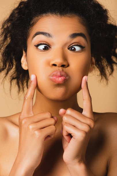 σγουρή Αφροαμερικανή γυναίκα με γυμνούς ώμους που αγγίζει και μουγκρίζει μάγουλα απομονωμένα σε μπεζ  - Φωτογραφία, εικόνα