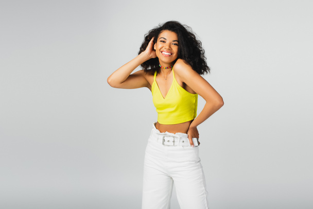 pleased african american woman in yellow crop top posing with hand on hip isolated on grey - Foto, afbeelding