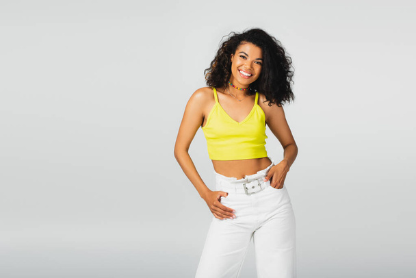 happy african american woman in yellow crop top and white pants posing with hand in pocket isolated on grey  - Fotoğraf, Görsel