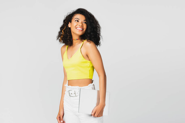 positive african american woman in yellow crop top holding laptop isolated on grey - Photo, image