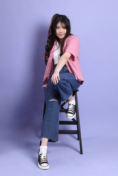 Милая молодая азиатская девушка в повседневной одежде сидит на фиолетовом фоне. - Фото, изображение