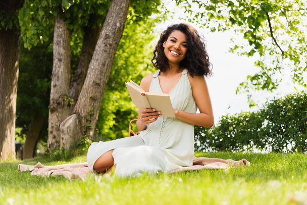 веселая африканская американка в платье держит книгу и сидит на одеяле в парке - Фото, изображение