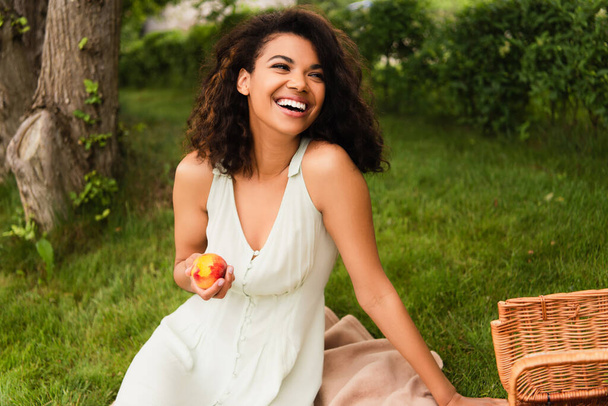 Веселая африканская американка в белом платье держит персик возле плетеной корзины в парке  - Фото, изображение