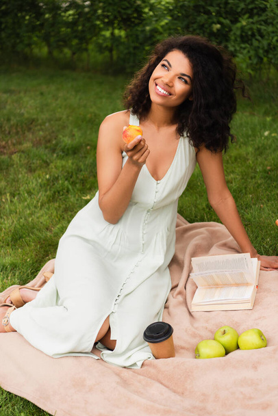 gelukkig en jong Afrikaans amerikaans vrouw in wit jurk holding perzik in park  - Foto, afbeelding