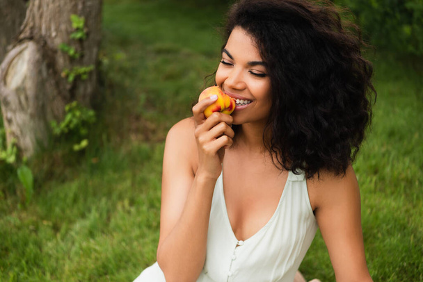 счастливая и молодая африканская американка в белом платье ест персик в парке  - Фото, изображение
