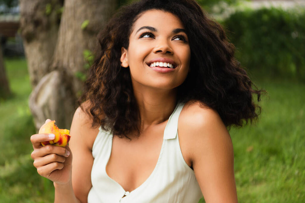 happy and young african american woman in white dress holding peach in park  - Photo, image