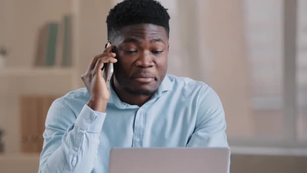 アフリカ人男性ビジネスマンの笑顔フリーランスマルチタスクワーカーは、ノートパソコン画面で携帯電話の外観に自宅のオフィストークに座るリモート通信ビジネスコールを提供コンピュータサービスを使用しています - 映像、動画