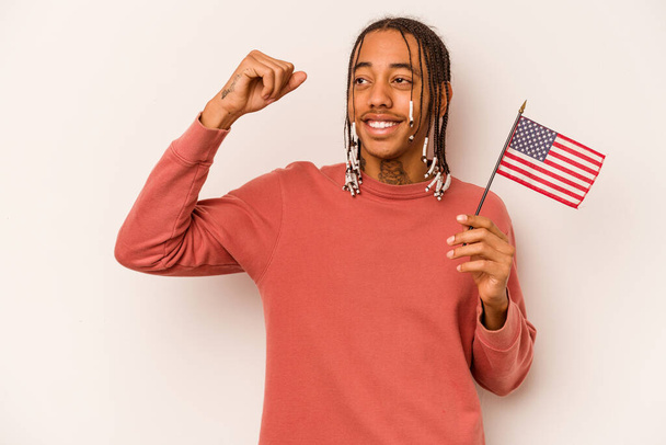 Молодой афроамериканец, держащий американский флаг на белом фоне, поднимающий кулак после победы, концепция победителя. - Фото, изображение