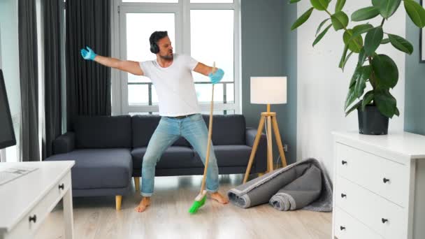 Hombre caucásico en auriculares limpiando la casa y divirtiéndose bailando con una escoba. Movimiento lento - Imágenes, Vídeo