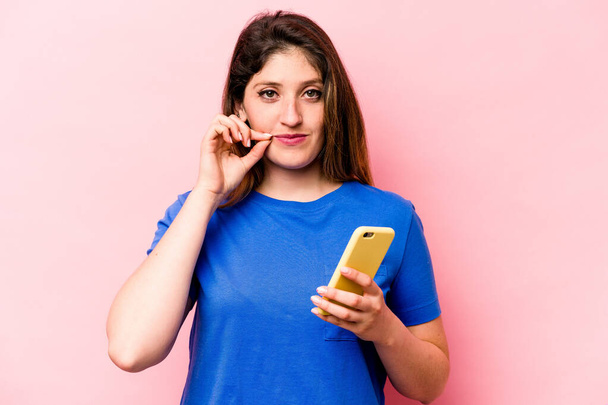 Young caucasian woman holding mobile phone isolated on pink background with fingers on lips keeping a secret. - Fotografie, Obrázek