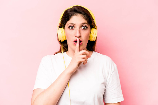 Young caucasian woman listening to music isolated on pink background keeping a secret or asking for silence. - Photo, image