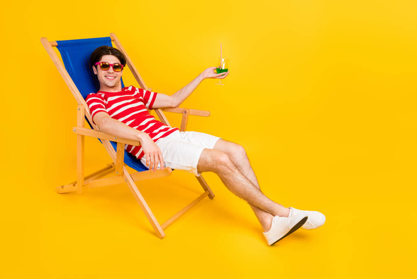 Photo of shiny cute young guy dressed red t-shirt smiling sitting lounge chair drinking cocktail isolated yellow color background. - Photo, image