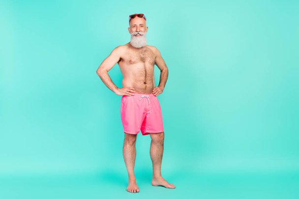 Фото красивого бородатого пенсионера мужчина руки бедра блестящая улыбка носить солнцезащитные очки розовые шорты изолированный бирюзовый фон. - Фото, изображение