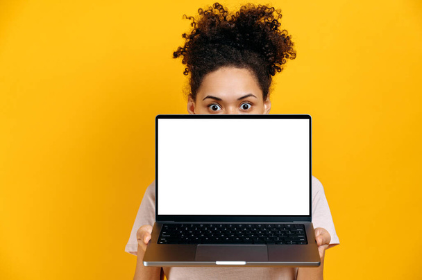 Amazed excited brunette african american girl peeking out from behind laptop, looks surprised at camera, stands on isolated orange background, holds an open laptop with blank white mock-up screen - Photo, Image