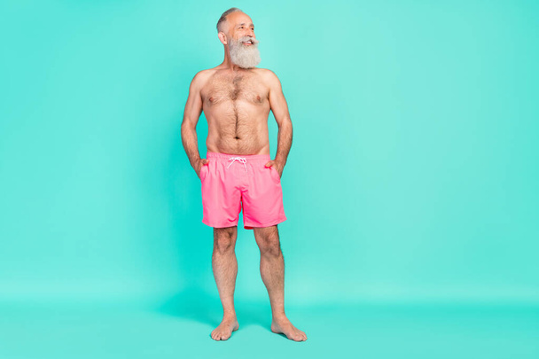 Photo of thoughtful masculine old man hands pockets look empty space wear pink shorts isolated teal color background. - Фото, изображение