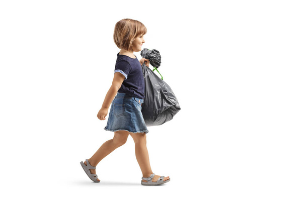 Full length profile shot of a little girl walking and carrying a waste bag isolated on white background - Photo, Image