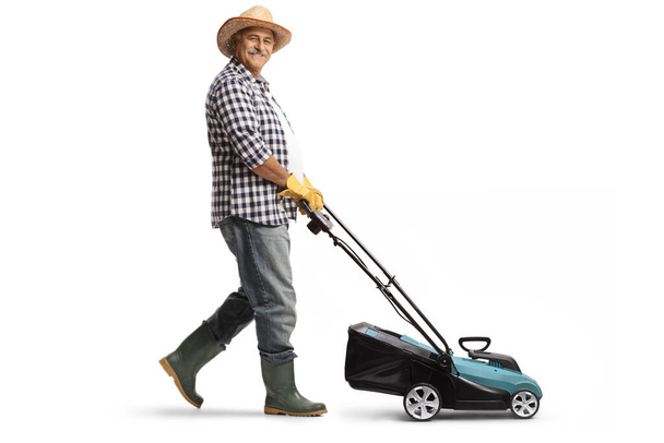Full length profile shot of a mature man with a straw hat and boots using a lawn mower isolated on white background - Fotó, kép
