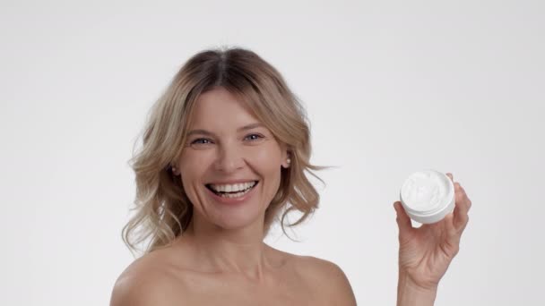 Luxury anti aging cosmetics. Close up portrait of happy beautiful middle aged lady demonstrating organic cream jar, smiling over white studio background, slow motion - Footage, Video