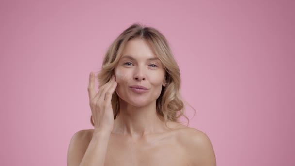 Beauty care. Close up portrait of beautiful mature lady applying moisturizing cream on face, pampering her skin, smiling to camera over pink studio background, slow motion - Footage, Video