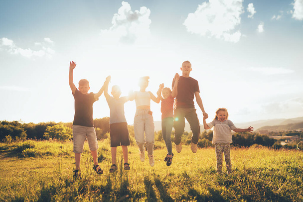 Six kids brothers and sisters teenagers and little kids funny jumping holding hands in hands on the green grass meadow with an evening sunset background light. Happiness and careless childhood concept. - Photo, Image