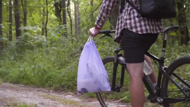 Ecologia, poluição, lixo e plástico descartados. Ciclista masculino fica com saco de lixo depois de coletar lixo plástico na floresta durante o ciclismo. Consciência de proibição de plástico, conceito de dia nacional de limpeza - Filmagem, Vídeo