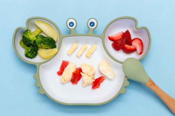 Baby Led weaning, blw. First toddler food. Soft eco plastic tableware with mixed vegetable. Healthy nutrition for kid, self feeding. Top view of silicone plate with spoon finger food for kid. - Photo, Image