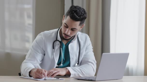 Hombre árabe concentrado médico se sienta en el gabinete de la clínica el uso de la computadora charlando distante en aplicación social médica dar consulta en línea escritura receta receta diagnosticar información de búsqueda - Metraje, vídeo