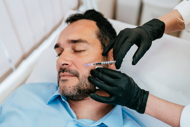 Handsome middle age bearded man is getting a rejuvenating facial injections at beauty clinic. The expert beautician is filling male wrinkles with botulinum toxin injections or hyaluronic acid fillers. - Valokuva, kuva