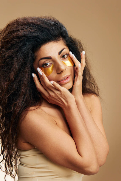 Hymyilevä houkutteleva Latinalaisen nainen soveltaa kulta hydrogeeli laastaria silmän alla nauttia ihonhoito poseeraa yli pastelli beige tausta. Kosmeettinen tuote mainos Natural kauneus käsite Studio muotokuva - Valokuva, kuva