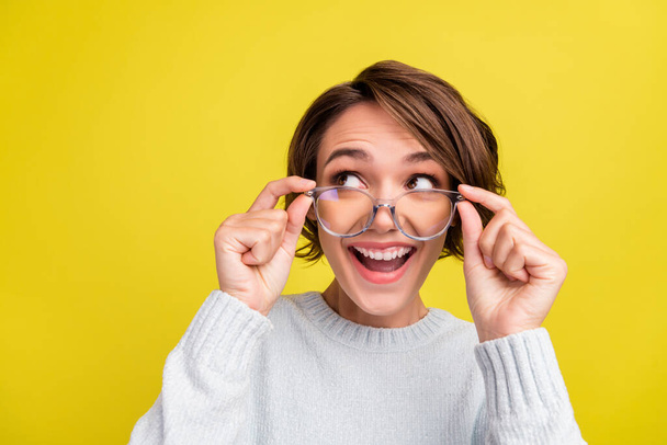 Photo of impressed young brunette lady hand eyewear look empty space wear blue sweater isolated on yellow color background. - 写真・画像