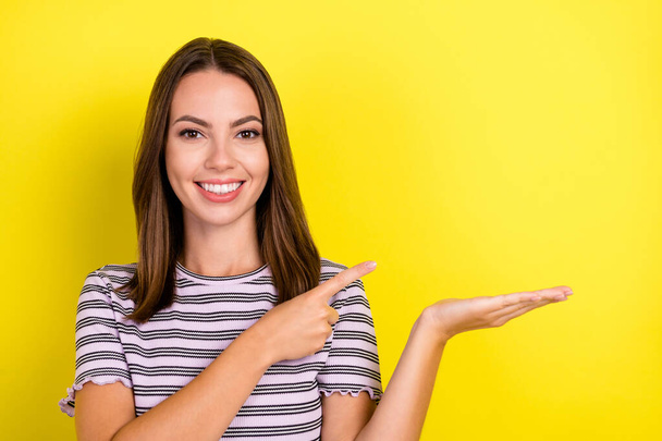 Photo of cute millennial brunette lady point hold empty space wear striped t-shirt isolated on yellow background. - Foto, Imagem