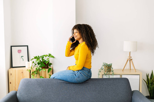 Jovem feliz usando telefone celular enquanto descansa em casa - Mulher alegre millennial falando no celular sentado no sofá - Foto, Imagem
