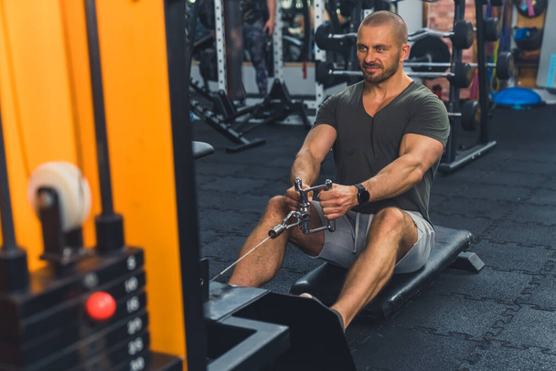 Front view of a good-looking sexy middle-aged guy with a beard doing exercise on a rowing machine in a modern well-equipped gym. . High quality photo - Foto, afbeelding