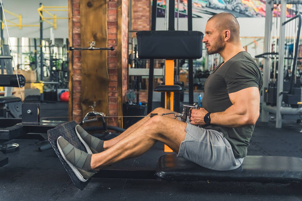 Bald tall european millenial man sitting on a bench tightening his muscles during an exercise on a rowing machine. High quality photo - Photo, image
