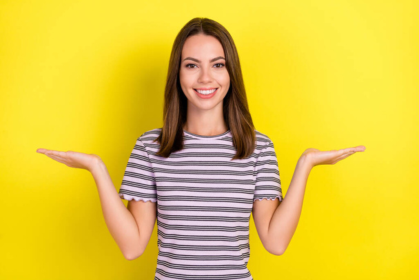 Photo of optimistic millennial brunette lady hold empty space wear striped t-shirt isolated on yellow background. - Фото, изображение