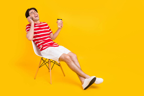 Full length photo of happy positive cheerful man talk phone hold coffee sit chair isolated on yellow color background. - Fotoğraf, Görsel