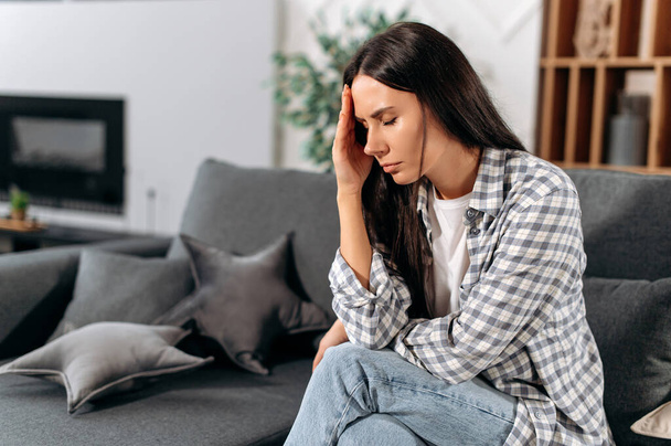 Headache, illness, personal experiences. Sad caucasian brunette woman, in casual clothes, sitting at home in the living room on the sofa, closed her eyes, experiencing stress, personal problems - Photo, Image